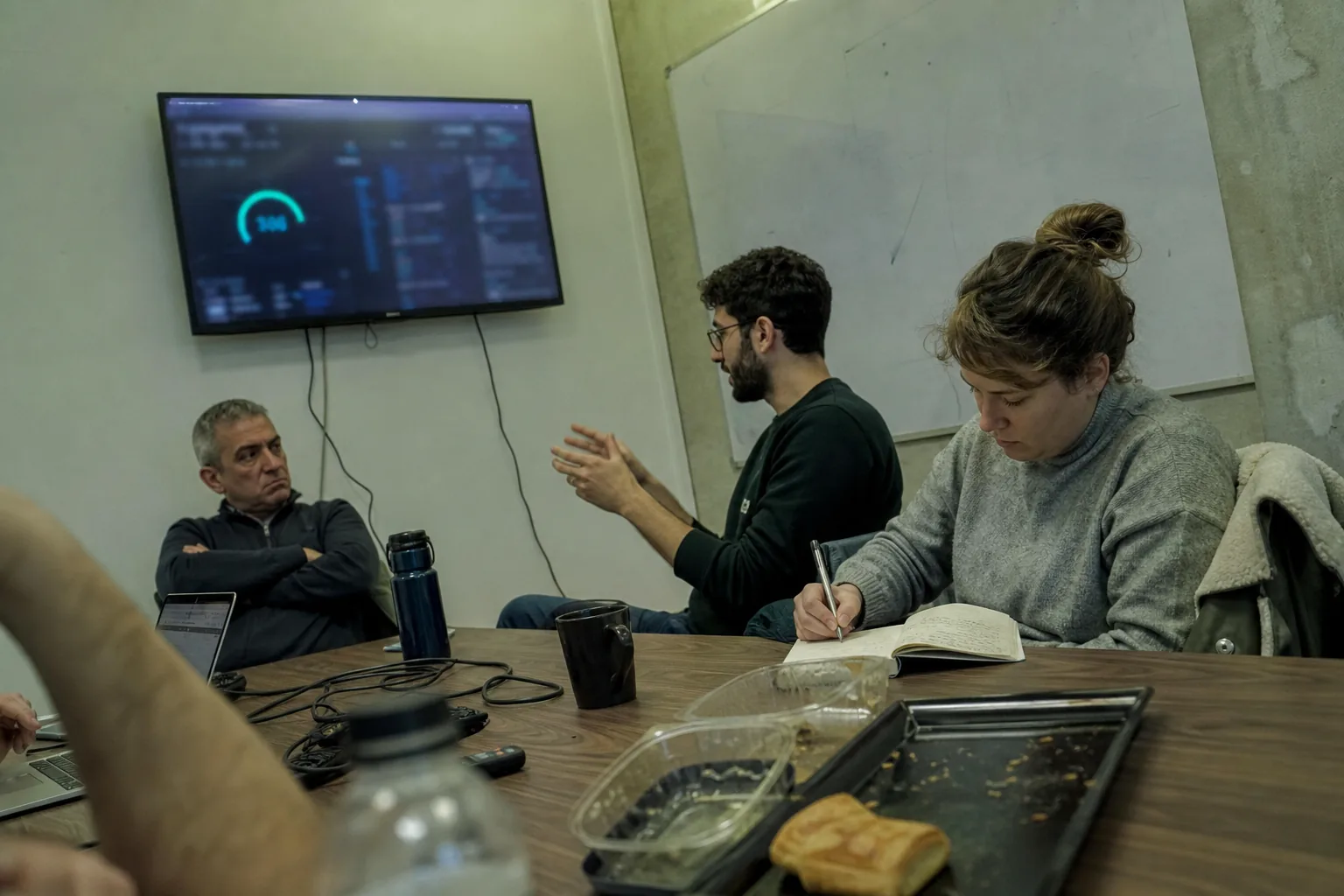 A Monday meeting, one person explaining a figure on the wall screen while another looks unconvinced