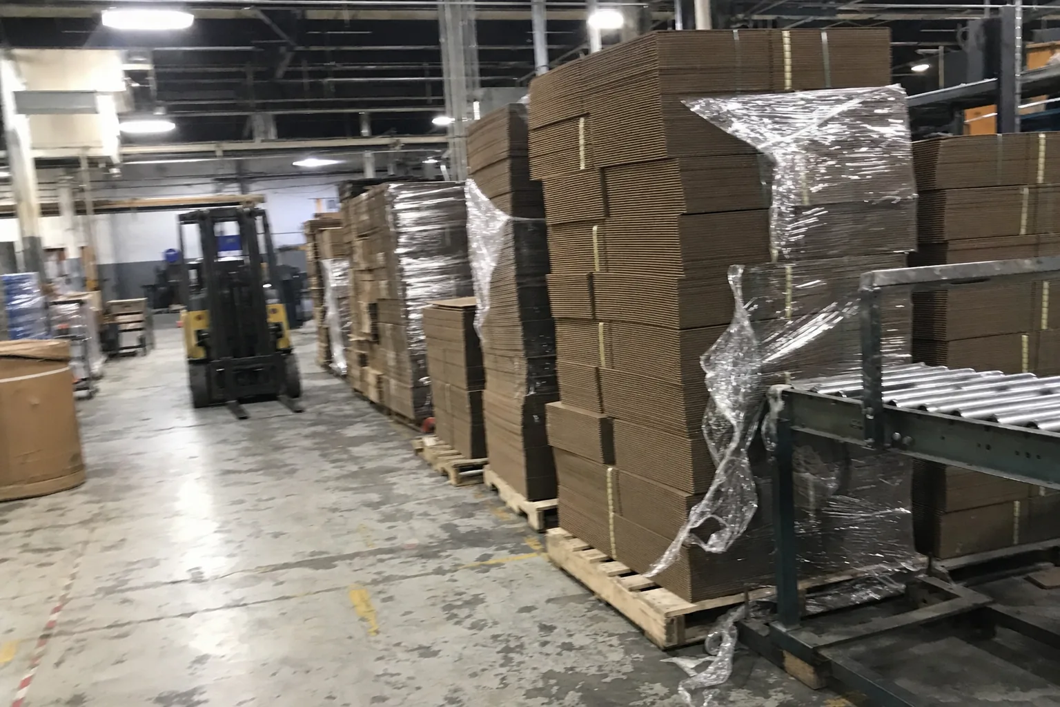 A worker in a hi-vis vest stacking flat corrugated sheets on a pallet, a forklift behind, in a box factory