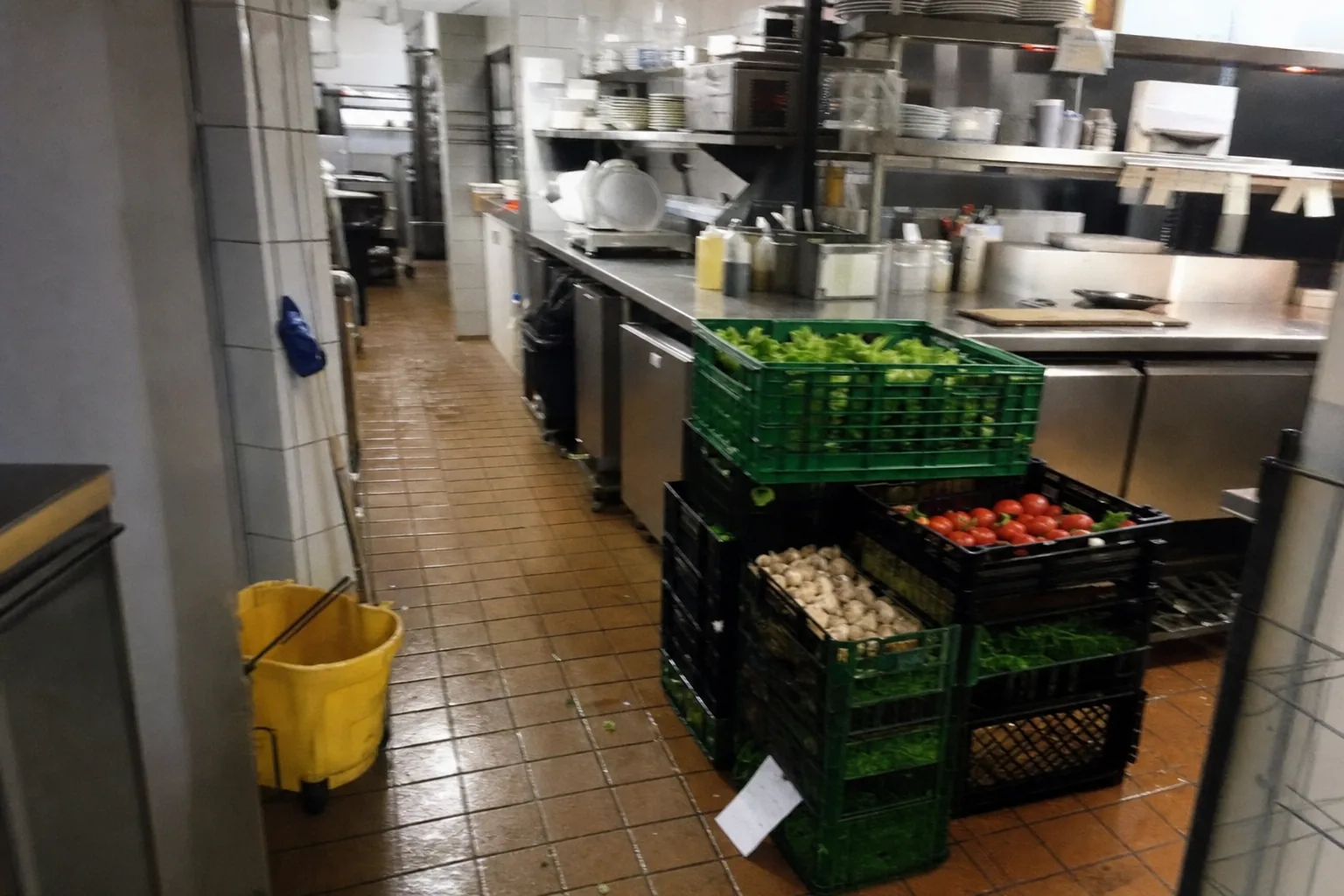 Crates of produce stacked on the floor of a restaurant kitchen before service, a mop bucket beside them